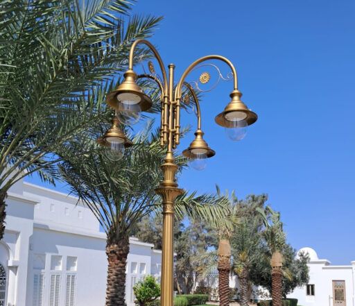 Reich verzierte Straßenlaterne mit goldenem Schirm und vier 04/Aries/A-Leuchten, vor einem Hintergrund aus Palmen und weißen Gebäuden an einem sonnigen Tag